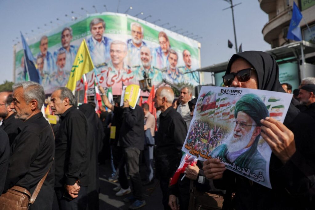 Warga mengikuti acara pemakaman di Teheran dengan latar belakang para petinggi militer Iran yang tewas dalam serangan Israel. (iranintl)