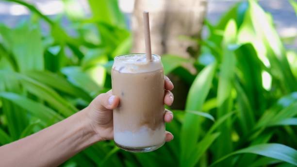 Segelas coconut milk coffee dingin dengan sedotan bambu dipegang tangan, latar daun hijau tropis alami.