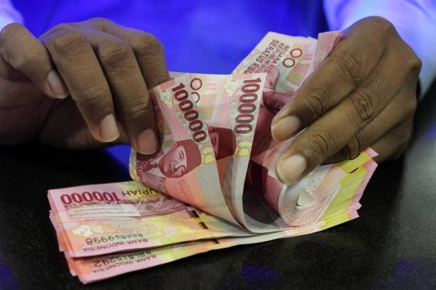 Tangan seseorang sedang menghitung lembaran uang rupiah pecahan seratus ribu di atas meja hitam.